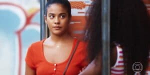 Jeniffer da novela Vai na Fé é uma mulher jovem, negra que está usando uma camiseta laranja e o cabelo está preso Jeniffer da novela Vai na Fé é uma mulher jovem, negra que está usando uma camiseta laranja e o cabelo está preso