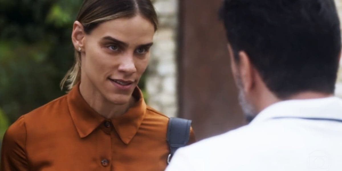 Sara usa camisa laranja e cabelo louro amarrado e conversa com Moretti de costa usando camisa branca na novela Travessia Sara usa camisa laranja e cabelo louro amarrado e conversa com Moretti de costa usando camisa branca na novela Travessia
